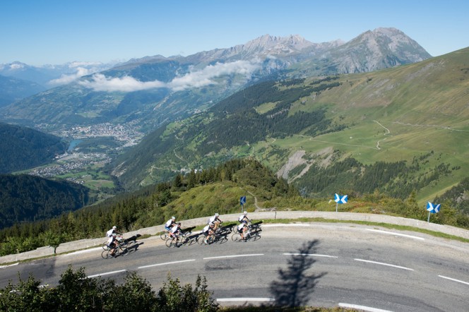 A large group of cyclists headed around a bend on road in the Alps.